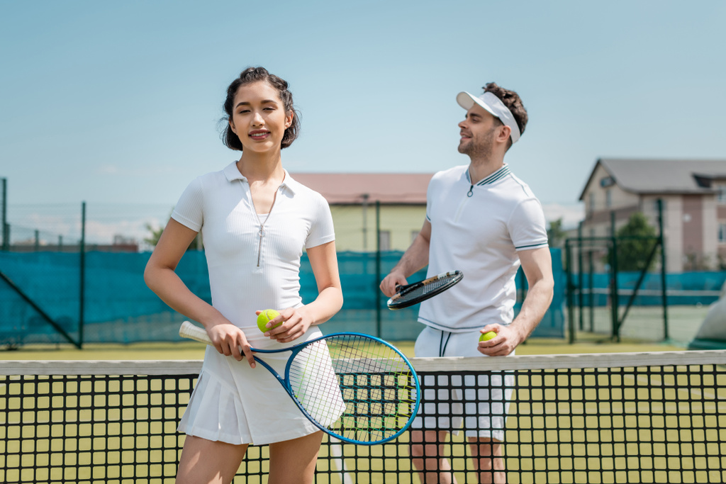 szczęśliwa kobieta w aktywnym ubraniu trzyma rakietę tenisową w pobliżu mężczyzny, tenisiści na korcie - Zdjęcie, obraz