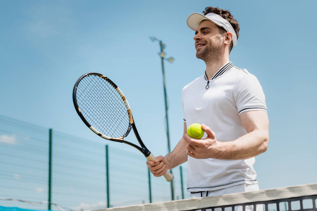 vrolijke tennisser in vizier cap holding racket en bal op het veld, fitness en motivatie, vreugde - Foto, afbeelding