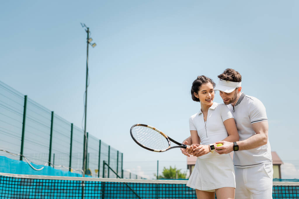 счастливый мужчина учит подругу играть в теннис на корте, держа рейнджеры и мяч, спорт и веселье - Фото, изображение
