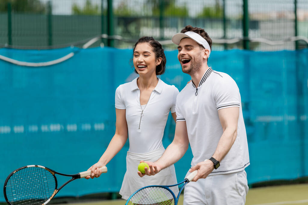 wesoła para w aktywnym ubraniu śmieje się na korcie tenisowym, gracze, rakiety i piłka, sport - Zdjęcie, obraz