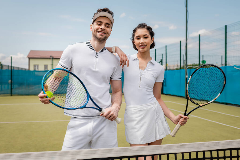 счастливая спортивная пара в активной одежде глядя на камеру на теннисном корте, хобби и спорт, лето - Фото, изображение