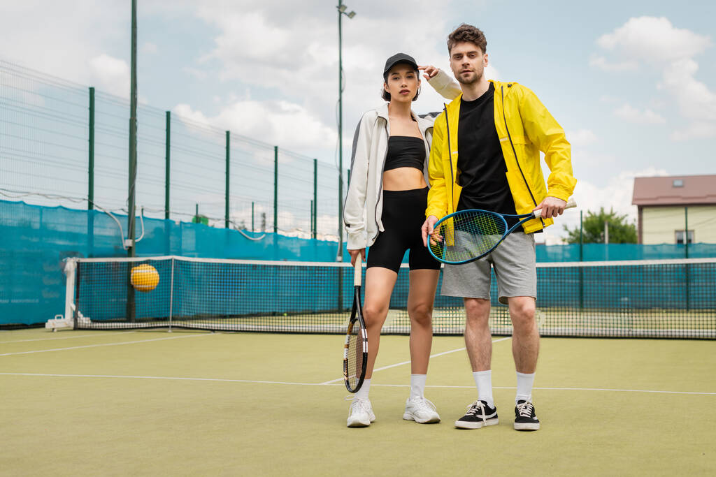 pareja de moda de pie en la cancha de tenis con raquetas, hombre y mujer en ropa deportiva, jugadores - Foto, Imagen