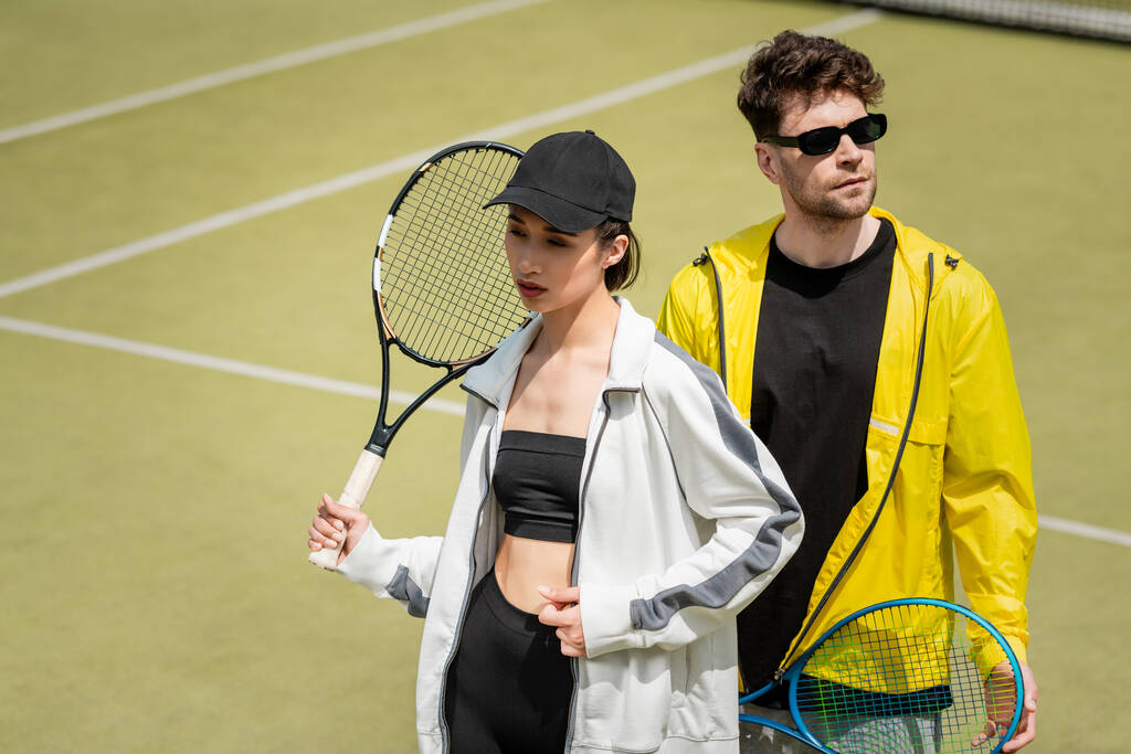 modisches sportliches Paar, Mann mit Sonnenbrille und Frau mit Schirmmütze mit Schlägern auf dem Tennisplatz - Foto, Bild