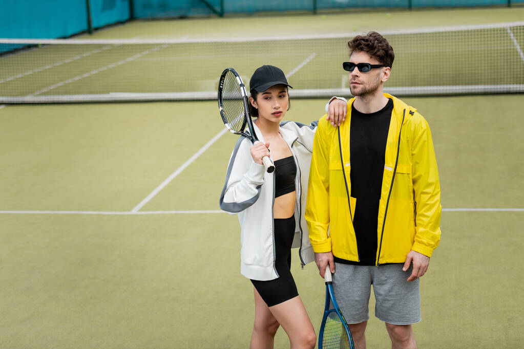 στύλ και αθλητισμός, αθλητικό ζευγάρι, άνδρας με γυαλιά ηλίου και γυναίκα με καπέλο που κρατά ρακέτες στο γήπεδο τένις - Φωτογραφία, εικόνα