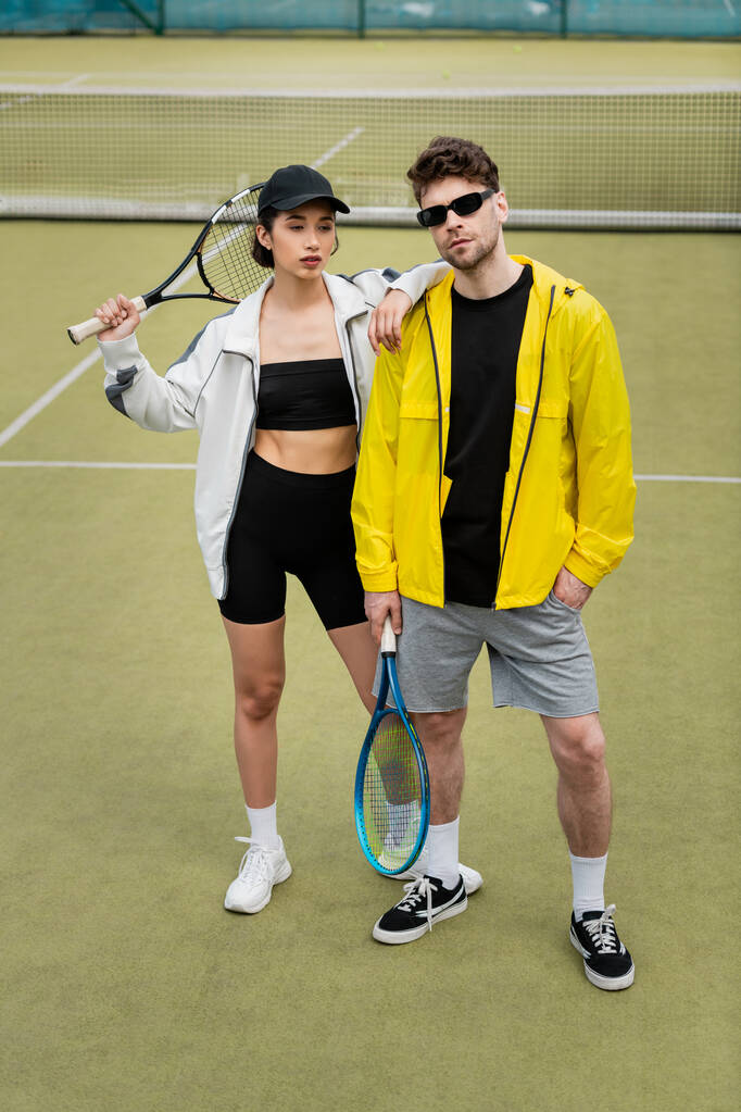 мода і спорт, спортсмен в сонцезахисних окулярах і жінка в кепці тримає ракетки на тенісному корті - Фото, зображення