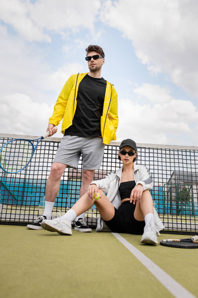 pareja deportiva, hombre y mujer en gafas de sol posando cerca de la red de tenis con raquetas, moda deportiva - Foto, Imagen