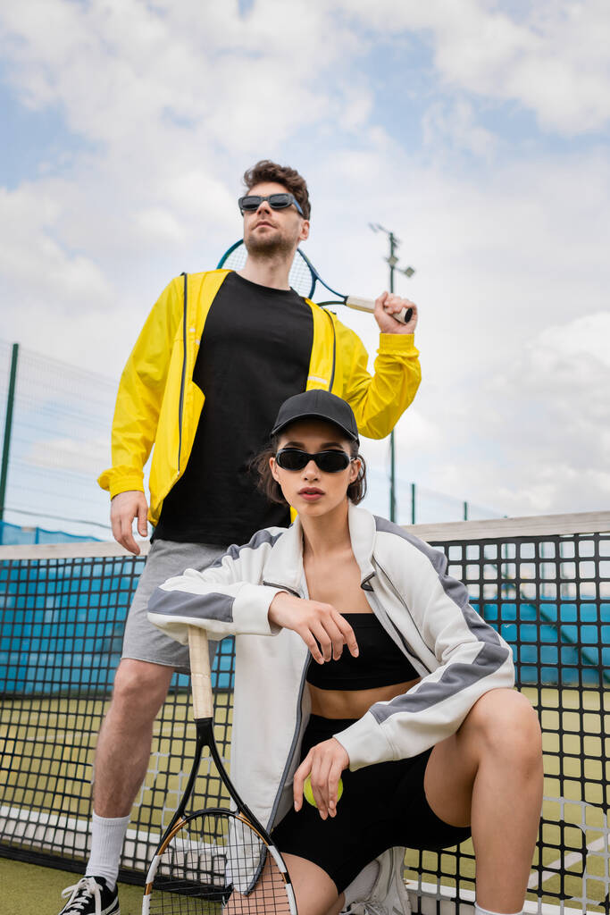 man en vrouw in zonnebril poseren in de buurt van tennis net met rackets, sportieve mode, sport als hobby, - Foto, afbeelding
