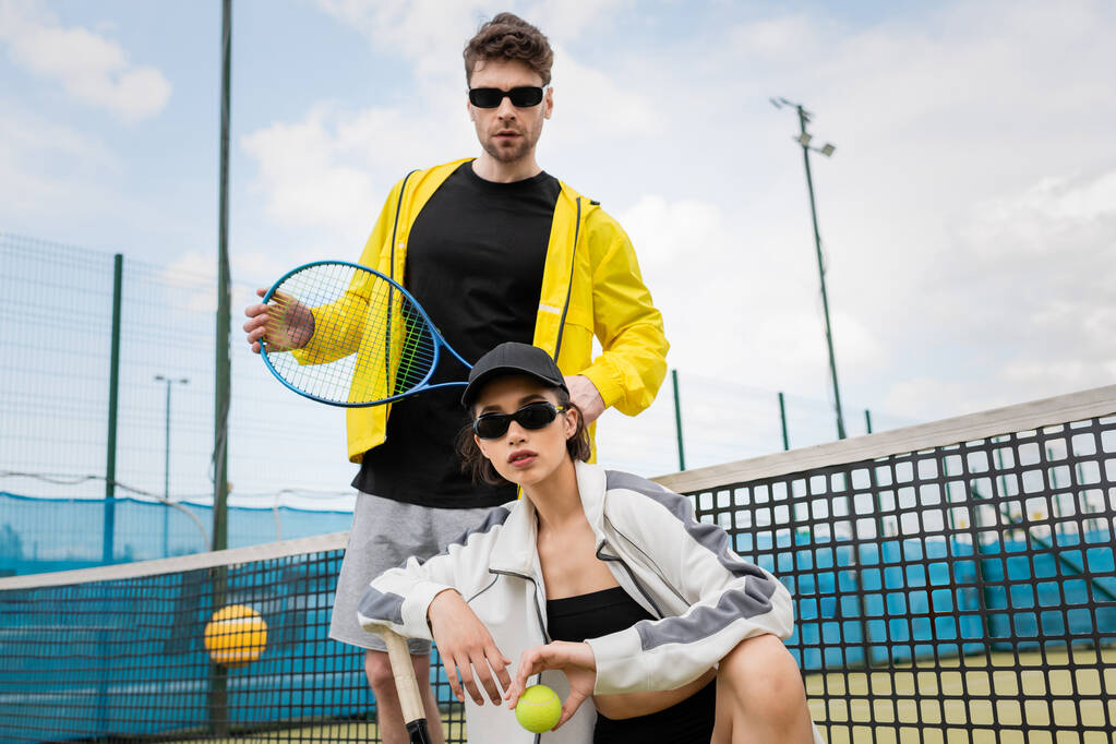 Güneş gözlüklü ve şapkalı bir kadın, adamın yanında poz veriyor, elinde top ve tenis raketi, etkin giyim tarzı. - Fotoğraf, Görsel