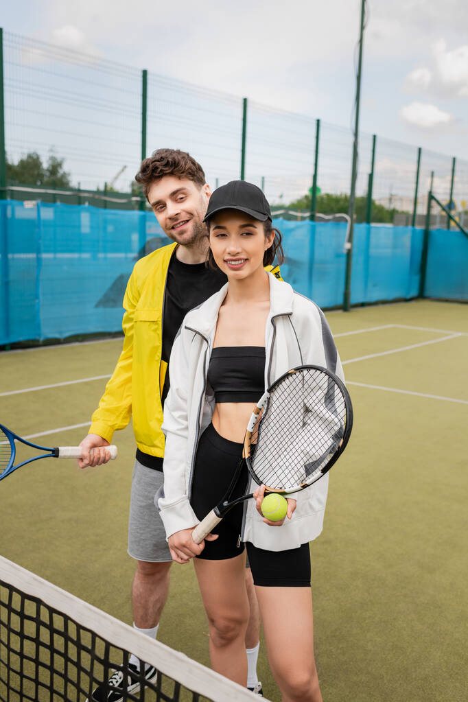 vrolijke man en vrouw in actieve dragen met tennisrackets en bal op het veld, op zoek naar camera - Foto, afbeelding