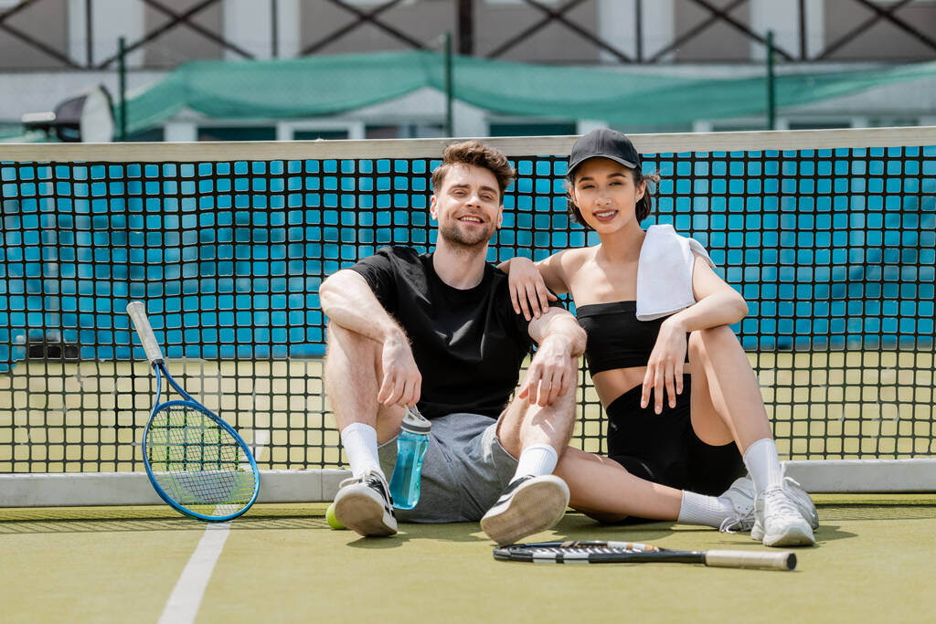 zdrowy tryb życia, szczęśliwy mężczyzna i kobieta w aktywnym ubraniu odpoczywający w pobliżu siatki tenisowej na korcie, rakiety - Zdjęcie, obraz