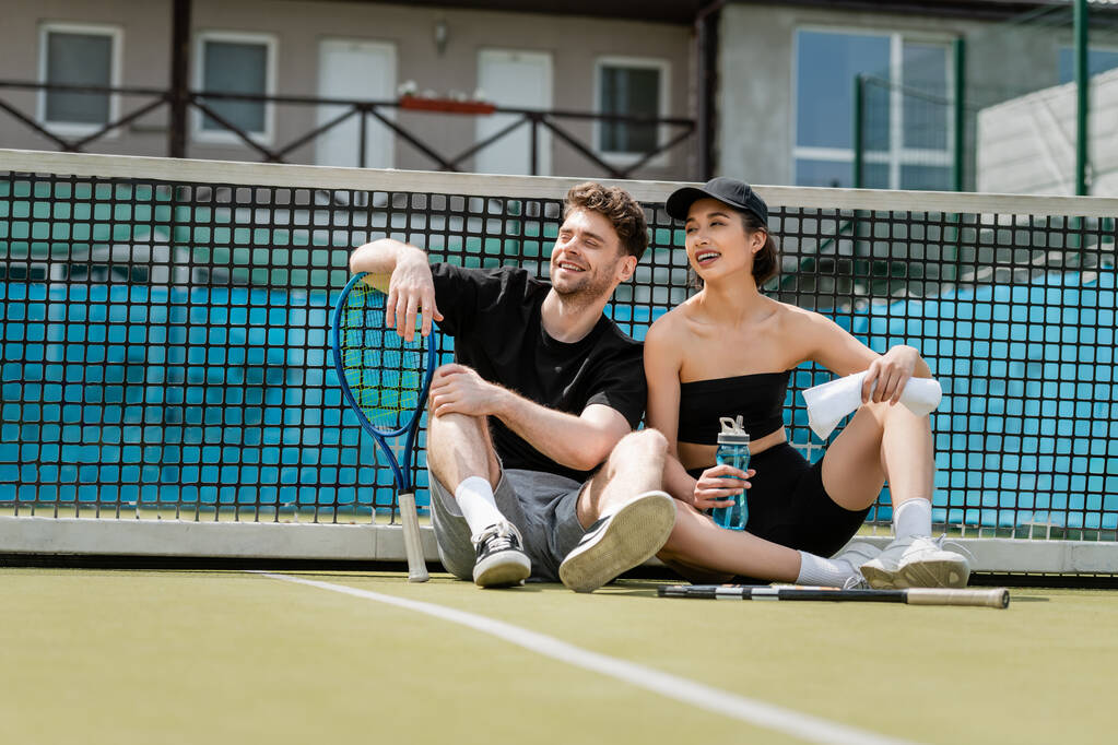 šťastný muž a žena ve sportovním oblečení odpočívá v blízkosti tenisové sítě s raketami na hřišti, zdravý životní styl - Fotografie, Obrázek