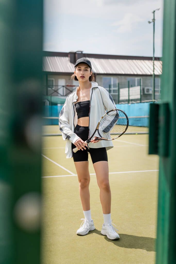 sportovní žena v aktivním oblečení a čepici stojící na tenisovém kurtu, držící raketu, dívající se do kamery - Fotografie, Obrázek