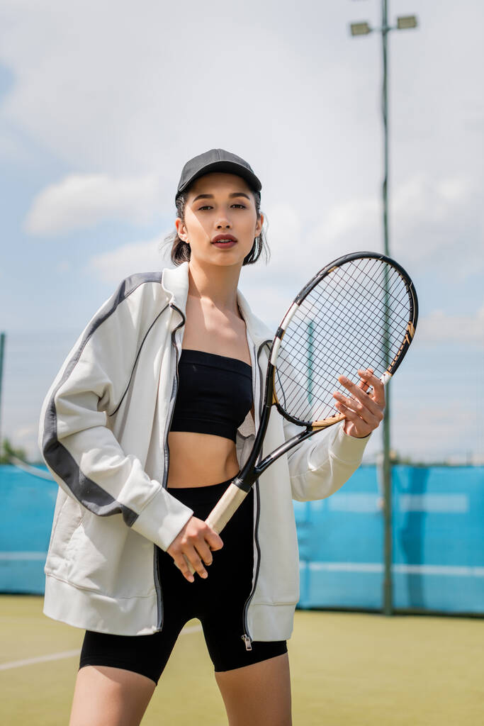 sportieve vrouw in actieve kleding en pet kijkend naar camera en tennisracket vasthoudend op het veld, motivatie - Foto, afbeelding