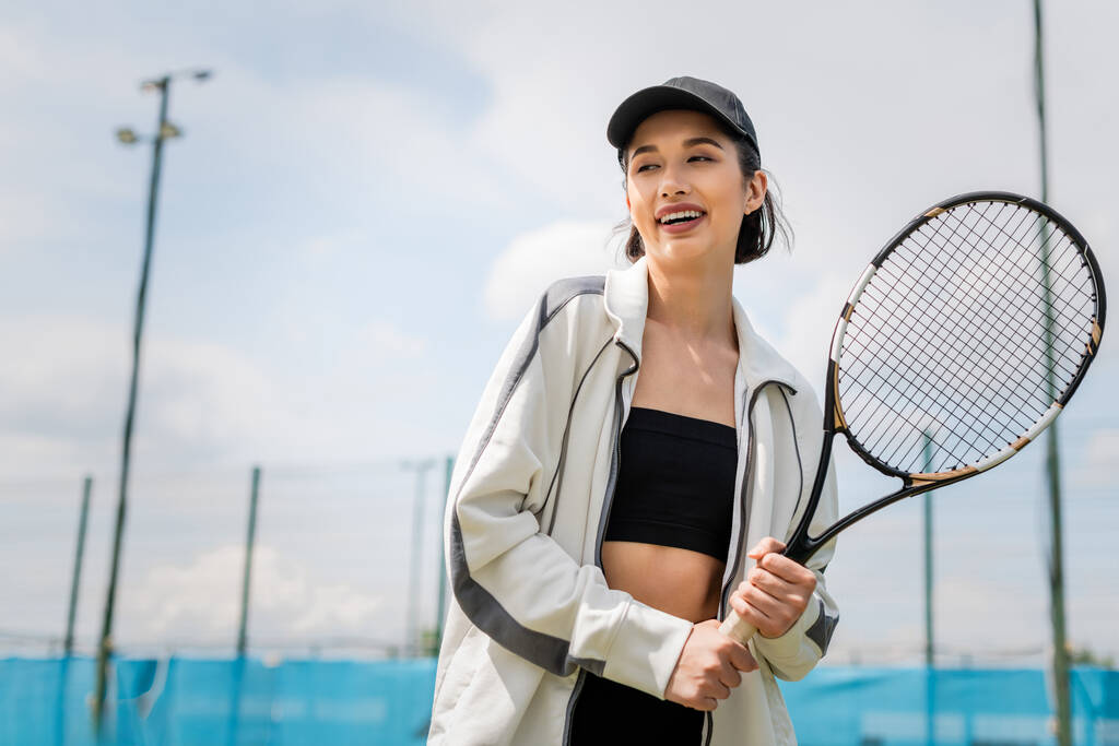 mulher feliz no desgaste ativo e boné segurando raquete de tênis na quadra, jogador de tênis feminino, motivação - Foto, Imagem