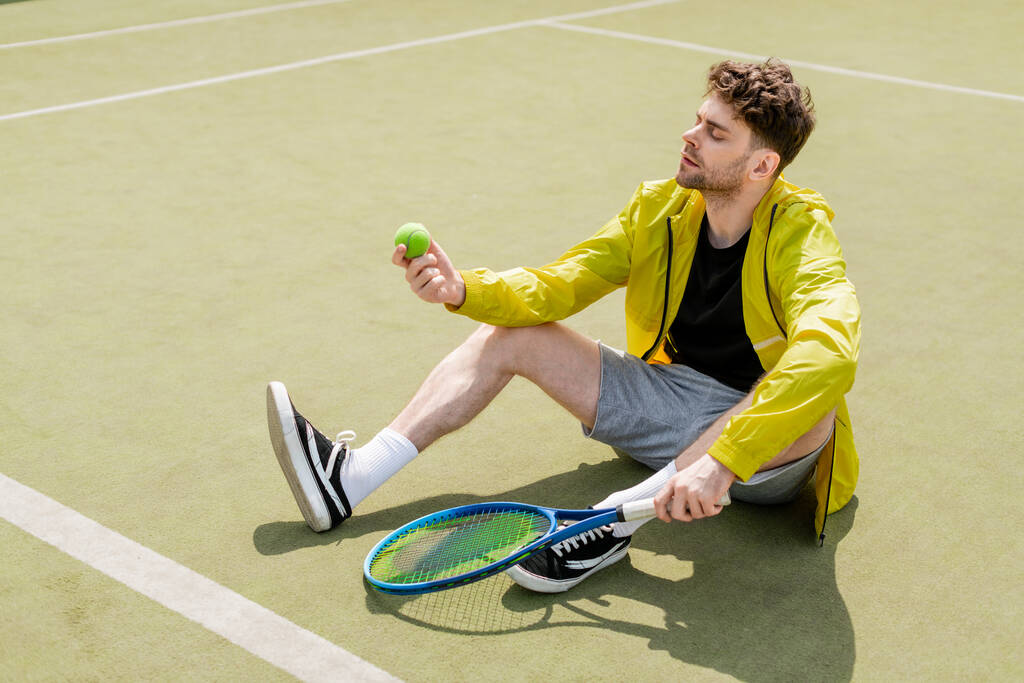 männlicher Tennisspieler sitzt auf dem Platz und hält Schläger in der Hand, schaut auf Tennisball, Mann in aktiver Kleidung - Foto, Bild
