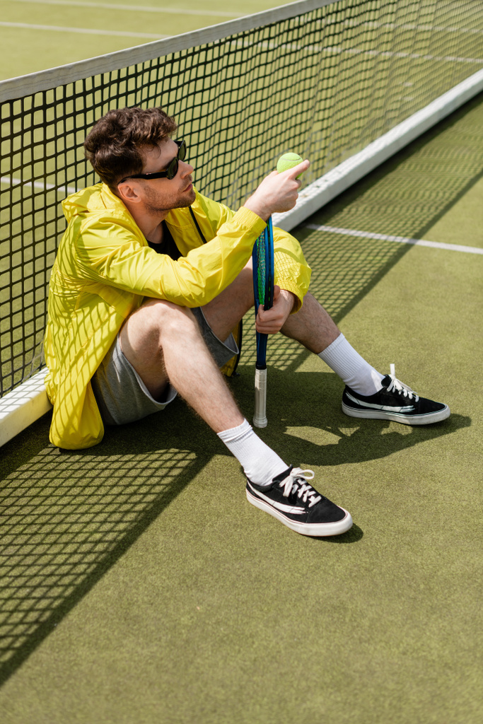 schöner Mann mit Sonnenbrille sitzt in der Nähe des Tennisnetzes, hält Ball und Schläger in der Hand, männlicher Tennisspieler - Foto, Bild