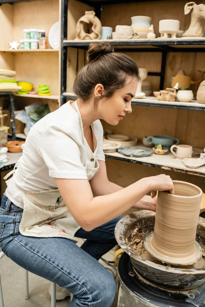 Side view of brunette craftswoman in apron working with clay on pottery wheel in studio in workshop - Photo, Image