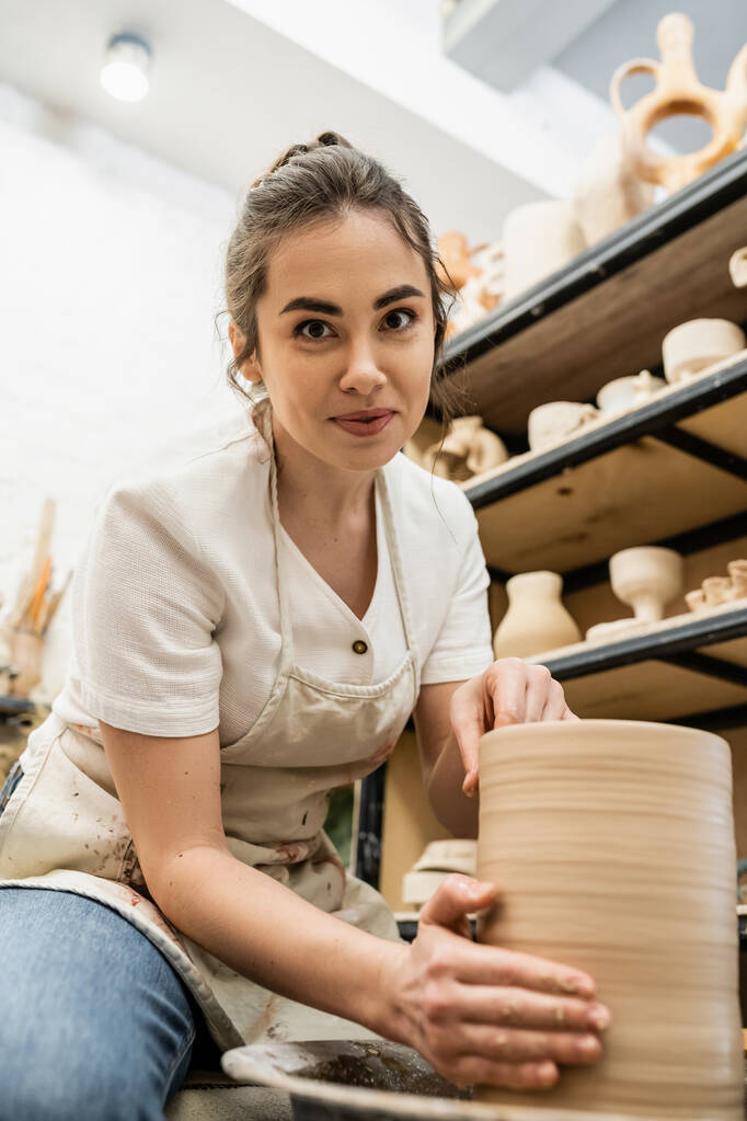 Brunette artisan en tablier regardant la caméra et la fabrication de vase d'argile sur roue de poterie dans l'atelier - Photo, image