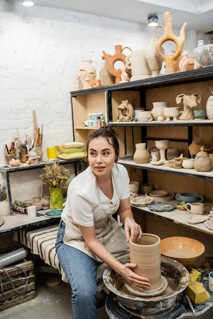 エプロンでブルネット女性職人 離れて見て,ワークショップで陶器の車輪に粘土花瓶を作る - 写真・画像