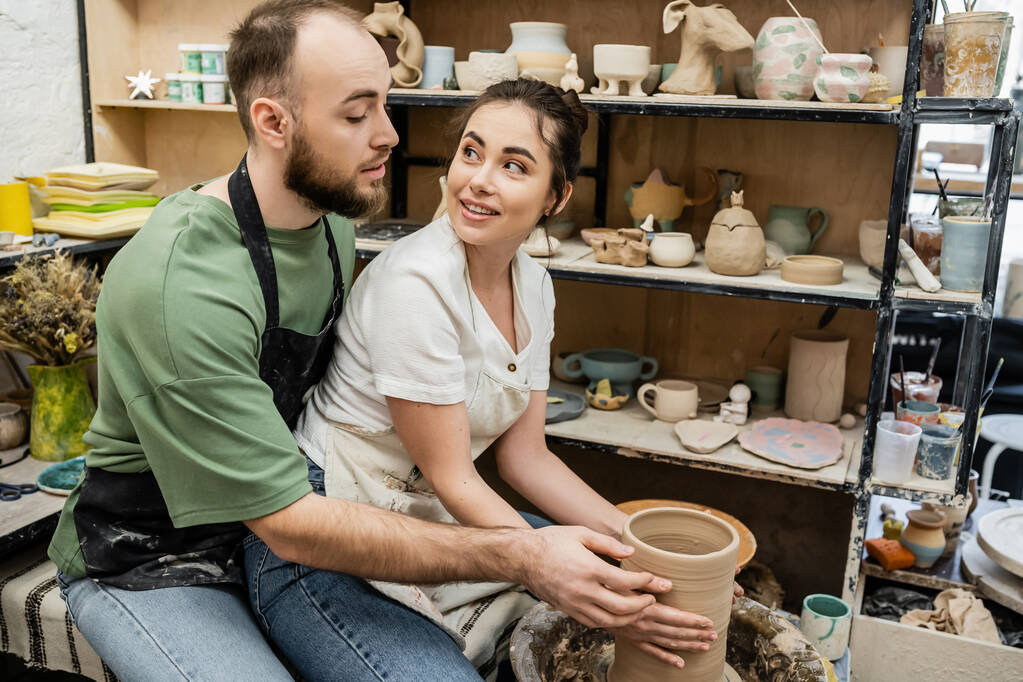 Uśmiechnięty rzemieślnik w fartuchu patrząc na chłopaka podczas wykonywania wazon gliniany na kole ceramiki w warsztacie - Zdjęcie, obraz