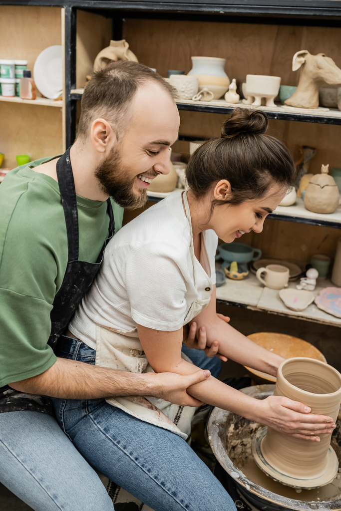 Vue latérale de joyeux artisans romantiques dans des tabliers faisant vase d'argile sur roue de poterie dans l'atelier - Photo, image