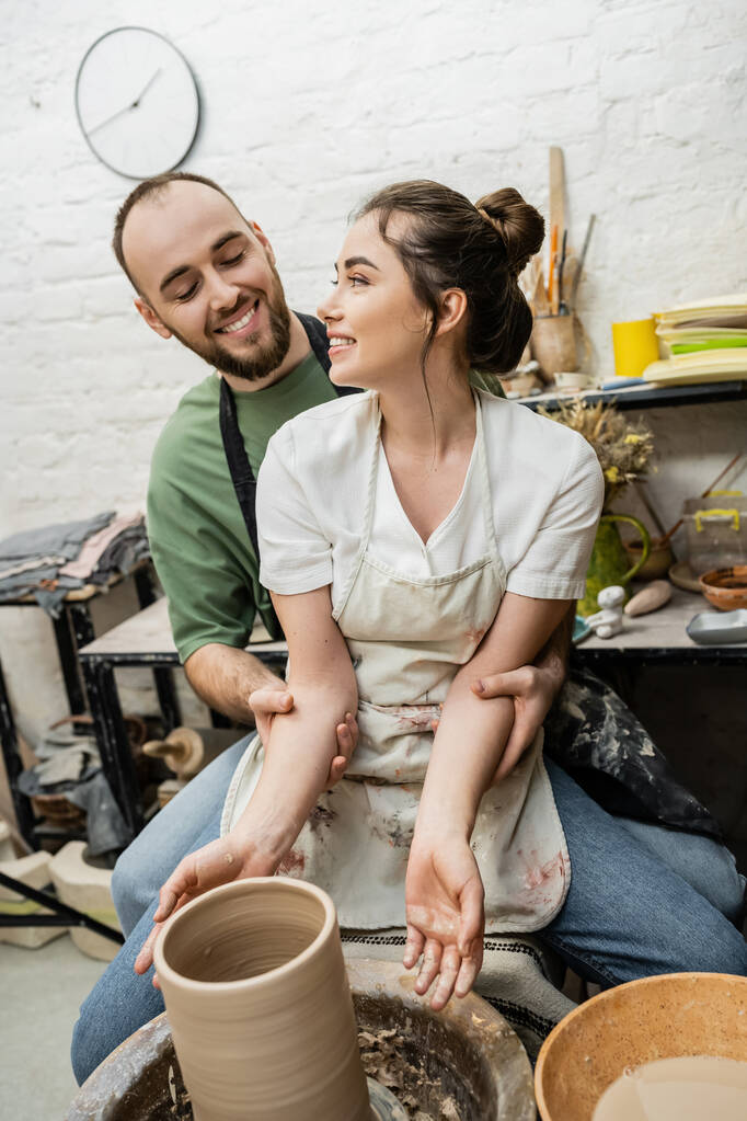Artisanat positif étreignant petite amie dans le tablier et faisant vase d'argile sur la roue de poterie en studio - Photo, image