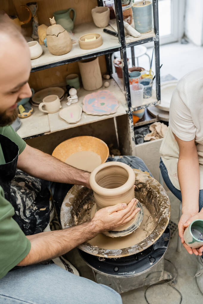 Vista de ángulo alto de la forma artesanal jarrón de arcilla en la rueda de cerámica cerca de la novia con taza en el estudio - Foto, imagen
