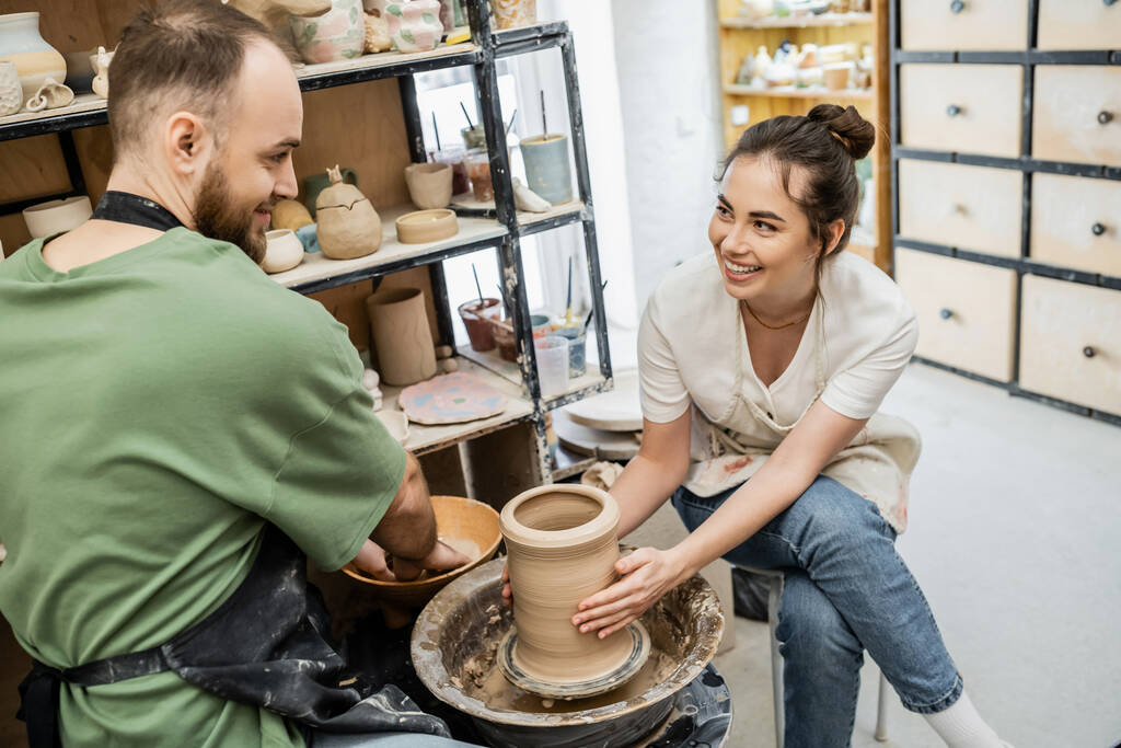 Artesana sonriente dando forma a jarrón de arcilla en la rueda de cerámica cerca de novio y tazón con agua en el estudio - Foto, imagen