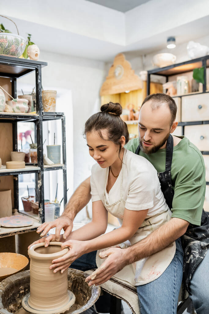 Bärtiger Handwerker im Gespräch mit fröhlicher Freundin beim Formen von Tonvasen auf Töpferscheibe im Keramikstudio - Foto, Bild