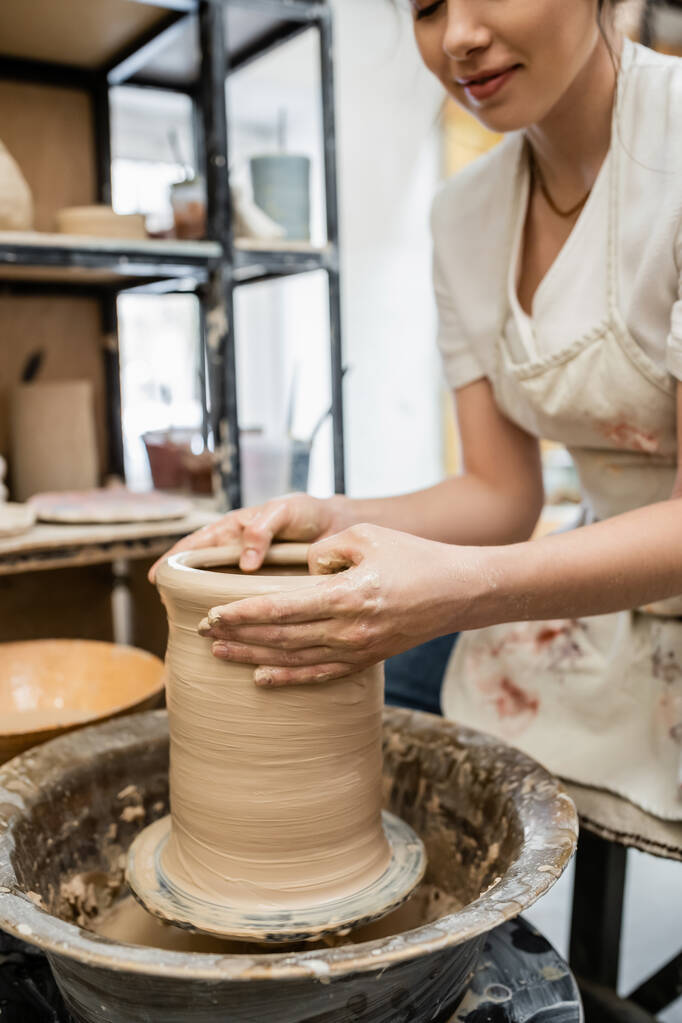 Cropped view of female potter in apron creating clay vase on pottery wheel in blurred workshop - Photo, Image