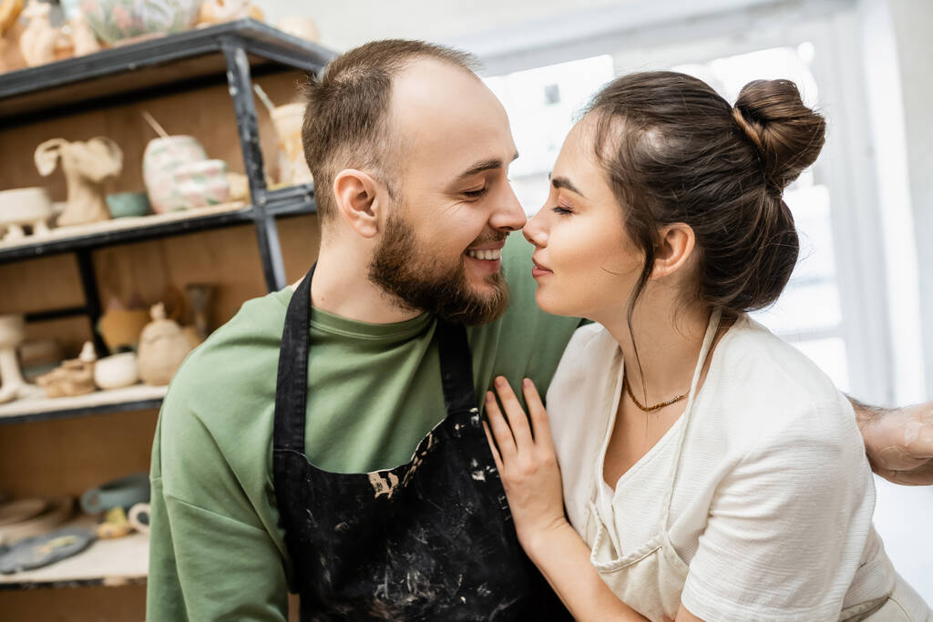 Side view of smiling sculptor in apron kissing brunette girlfriend in blurred ceramic workshop - Photo, Image