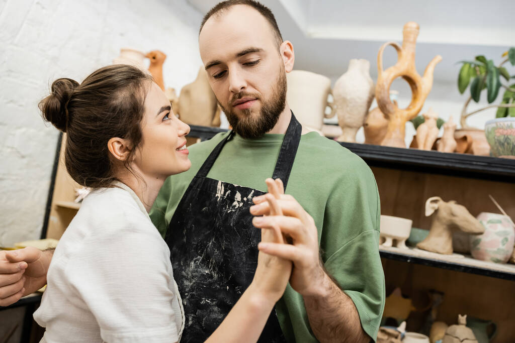 Бородатый скульптор в фартуке держит за руку улыбающуюся девушку во время работы в керамической мастерской - Фото, изображение