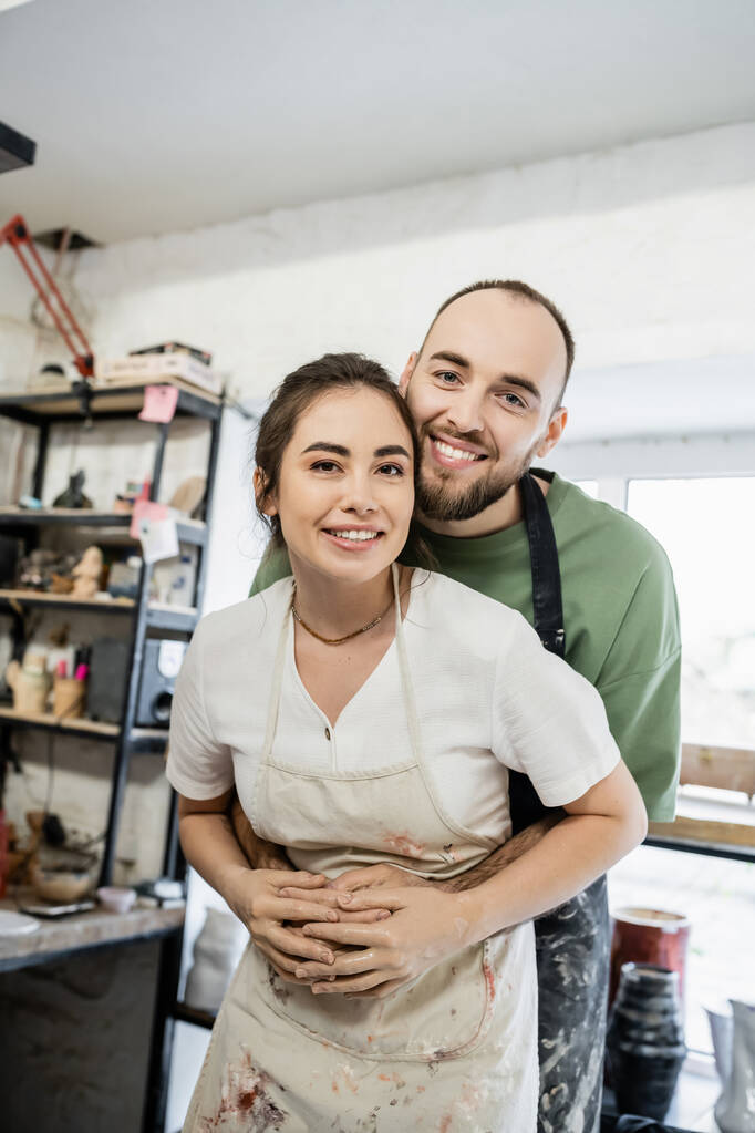 Positive bearded craftsman hugging girlfriend in apron and looking at camera in ceramic workshop - Photo, Image