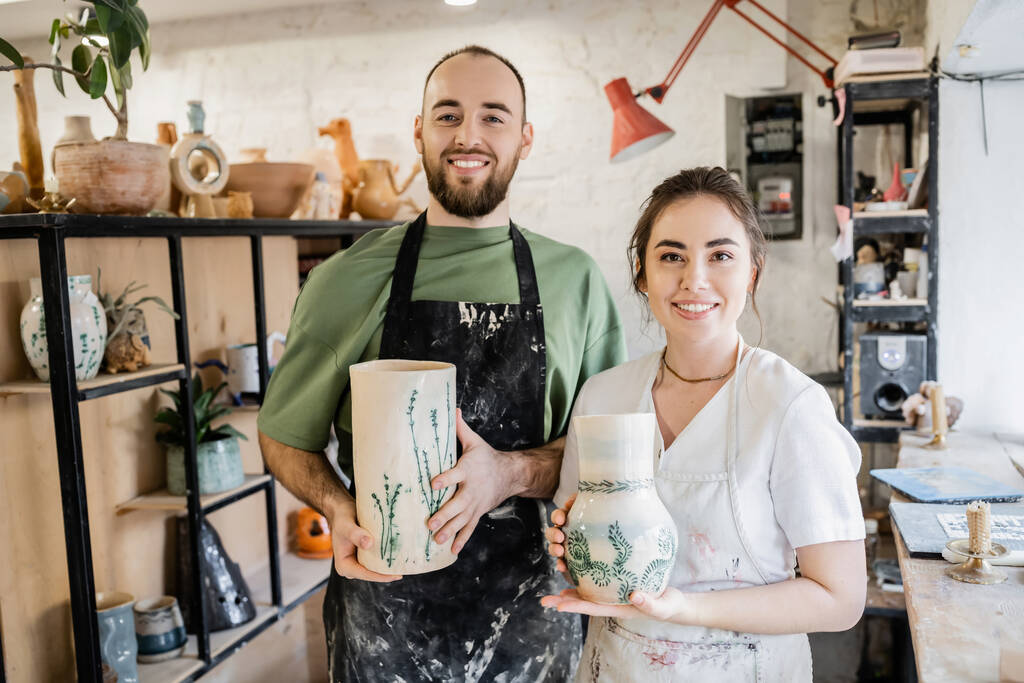 Pareja sonriente de artesanos en delantales sosteniendo jarrones de barro y mirando a la cámara en taller de cerámica - Foto, imagen