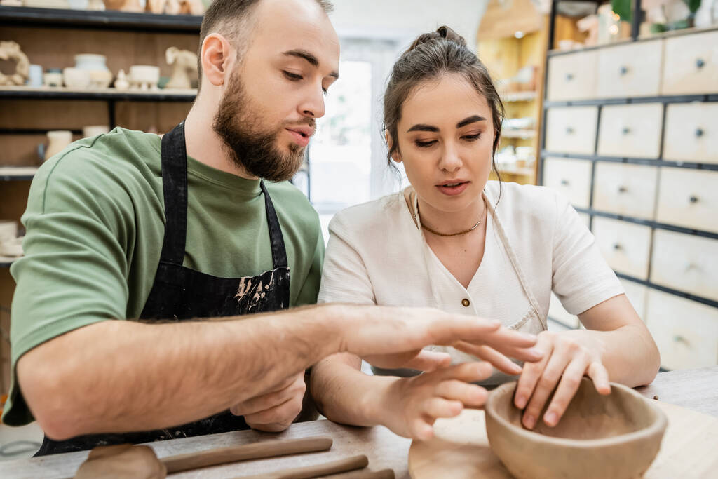 Bärtiger Handwerker in Schürze formt Tonschale und spricht mit Freundin in Keramik-Werkstatt - Foto, Bild