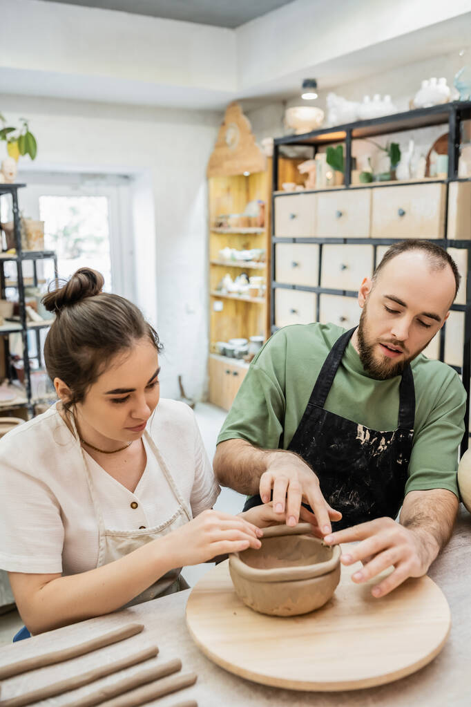 Пара гончаров в фартуках, формирующих глиняную чашу вместе во время работы в керамической мастерской - Фото, изображение