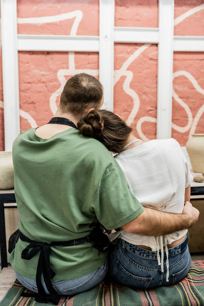 Πλάτη άποψη του τεχνίτη στην ποδιά αγκαλιάζει φίλη ενώ κάθεται κοντά σε πηλό στο στούντιο κεραμικής - Φωτογραφία, εικόνα