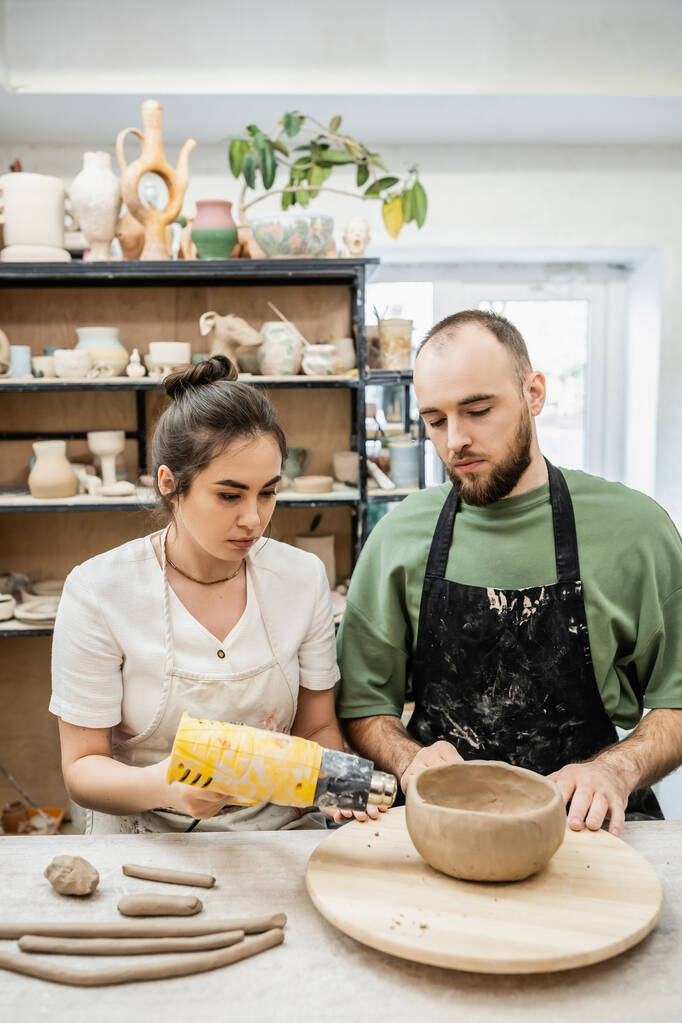 Couple d'artisans dans des tabliers cuvette en argile séchante avec pistolet à chaleur sur planche en bois dans un atelier de céramique - Photo, image
