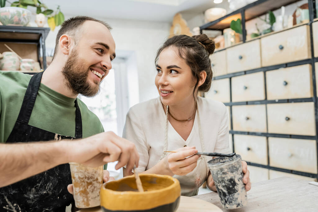 Lächelnde romantische Kunsthandwerker in Schürzen im Gespräch und Färbung Tonschale in Keramik-Werkstatt im Hintergrund - Foto, Bild