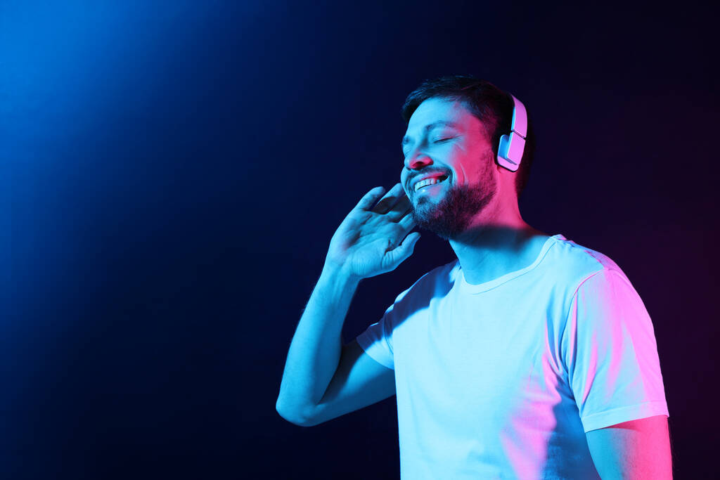 Szczęśliwy człowiek w słuchawkach cieszący się muzyką w neonach na ciemnoniebieskim tle. Miejsce na tekst - Zdjęcie, obraz