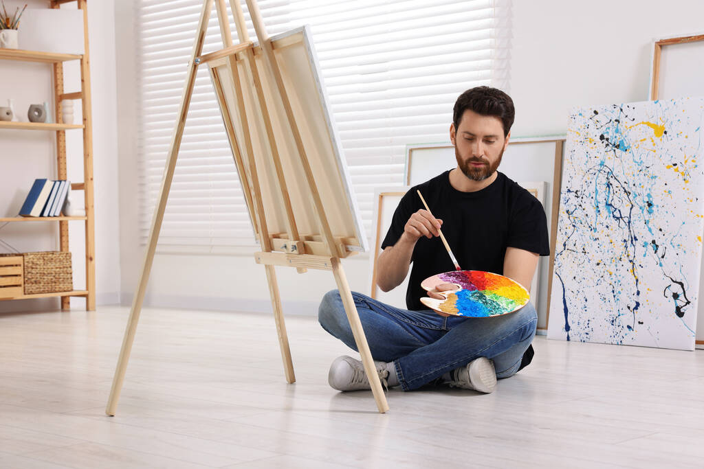 Man met penseel en palet schilderen in studio. Ezel gebruiken om doek vast te houden - Foto, afbeelding