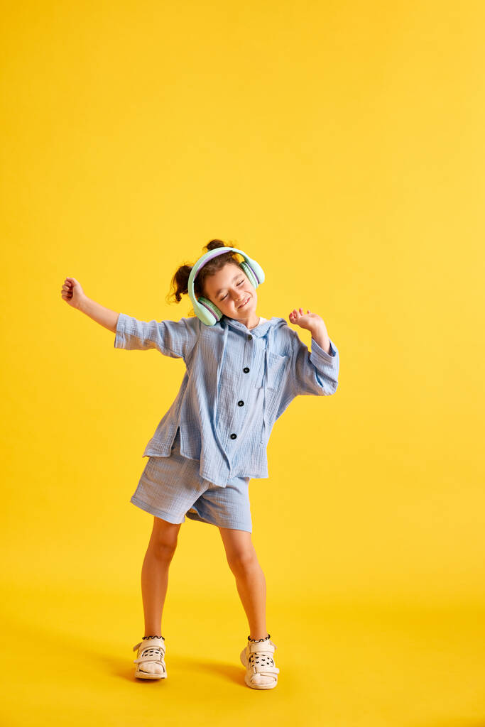 Full-length portrait of beautiful little child, girl listening to music in headphones and dancing against yellow studio background. Concept of emotions, childhood, education, fashion, lifestyle, ad - Photo, Image