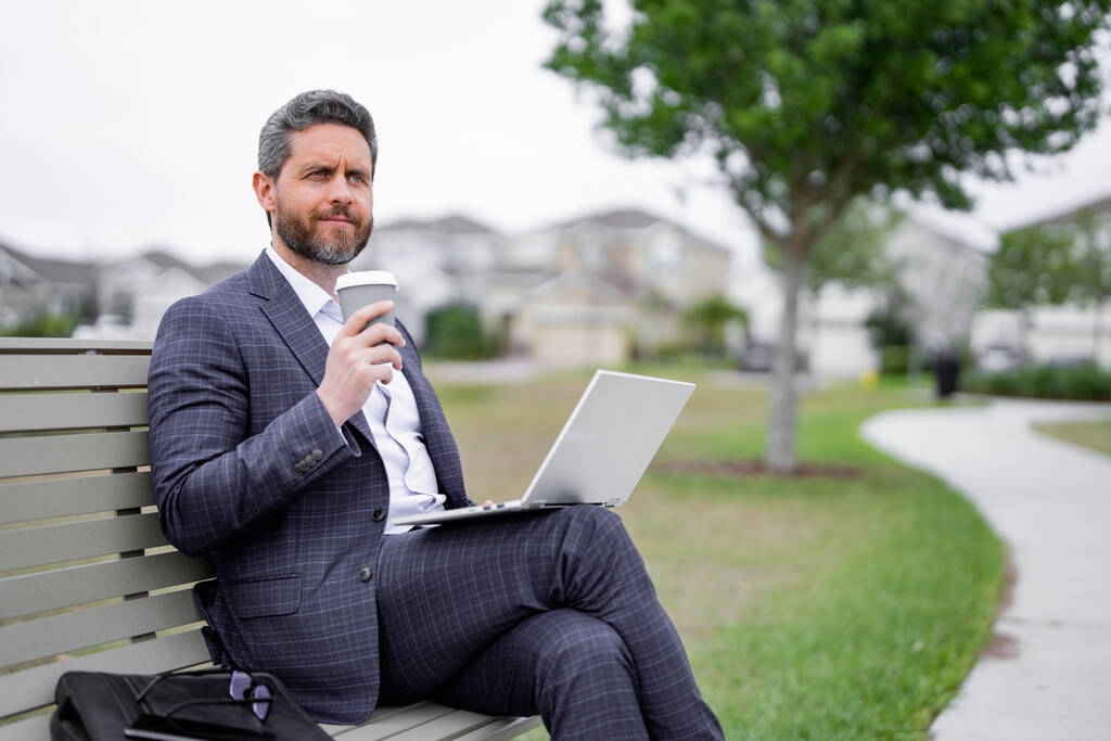 Üzletember ül a padon a parkban, laptopon dolgozik, kávét iszik. Üzletember online munka laptopról. Az üzletember pihen és híreket olvas az amerikai negyedben. Az üzletember a padon pihen. - Fotó, kép