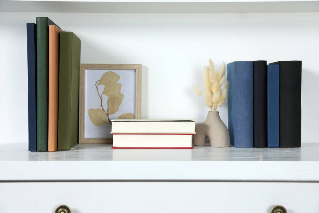 Книги в твердом переплете, картины и вазы с сухими цветами на белом комоде - Фото, изображение