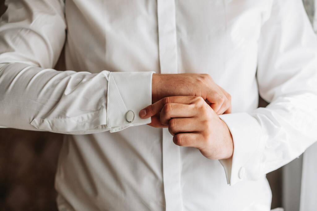 Portret van een modieuze man die stijlvolle manchetknopen vastmaakt op de mouwen van zijn witte shirt. Wedding day concept. - Foto, afbeelding