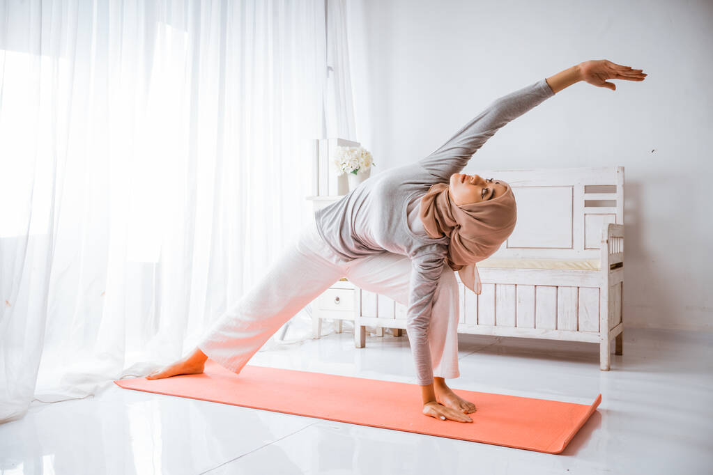 asiático musulmán mujer instructor usando hijab haciendo yoga pilates pose tutorial en naranja colchón en un habitación - Foto, imagen