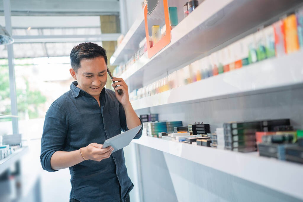 Erkek esnaf, telefon ederken ve dijital tablete bakarken mağazanın rafının yanında duruyor. - Fotoğraf, Görsel