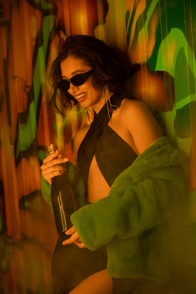 Femme brune positive en lunettes de soleil et robe tenant champagne près des graffitis dans la boîte de nuit - Photo, image