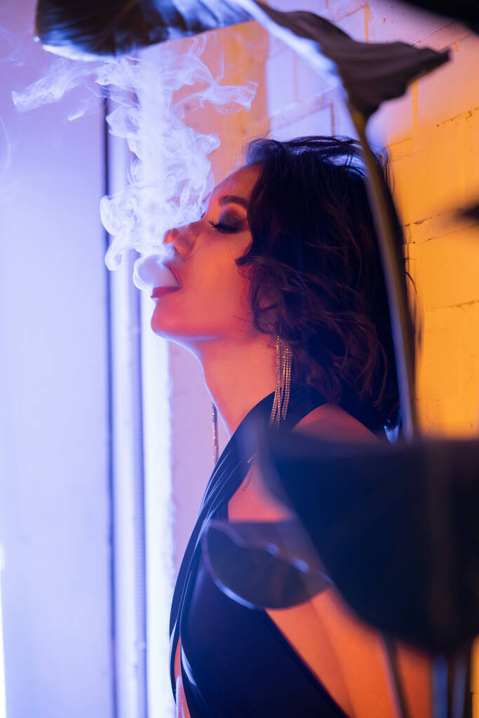 Trendy young brunette asian woman in dress exhaling smoke in neon light in night club - Photo, Image