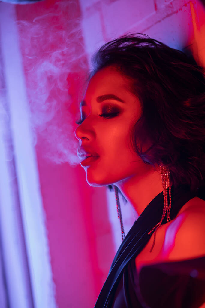 Módní mladá asijská žena s make-up vydechující kouř, zatímco stojí v neonovém světle v nočním klubu - Fotografie, Obrázek